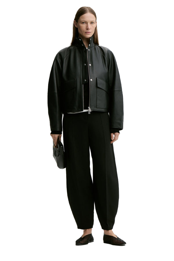 Front view of a woman wearing By Malene Birger jacket in black with patents, zipper and pockets, paired with black barrel-fit trousers, black top and a pair of black flats, shown against a plain white background.