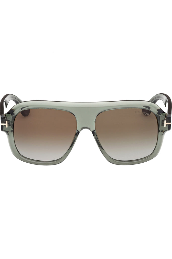 Tom Ford mask sunglasses in brown with translucent green frame and metal T details, shown against a plaid white background.