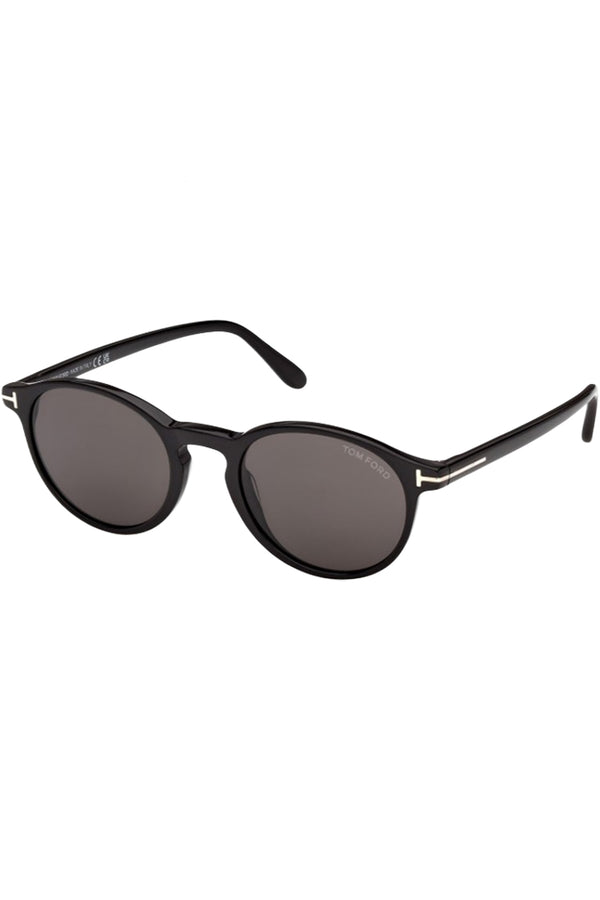 Rotated view of Tom Ford oval sunglasses in gray with black frame and metal T details, shown against a plaid white background.