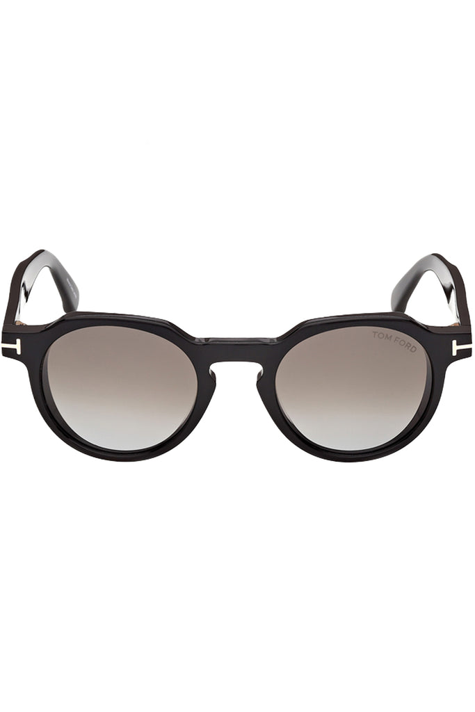 Tom Ford oval sunglasses in gray with black frame and metal T details, shown against a plaid white background.
