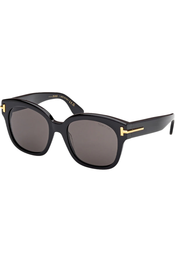 Rotated view of Tom Ford oval sunglasses in gray with black frame and gold T details, shown against a plaid white background.