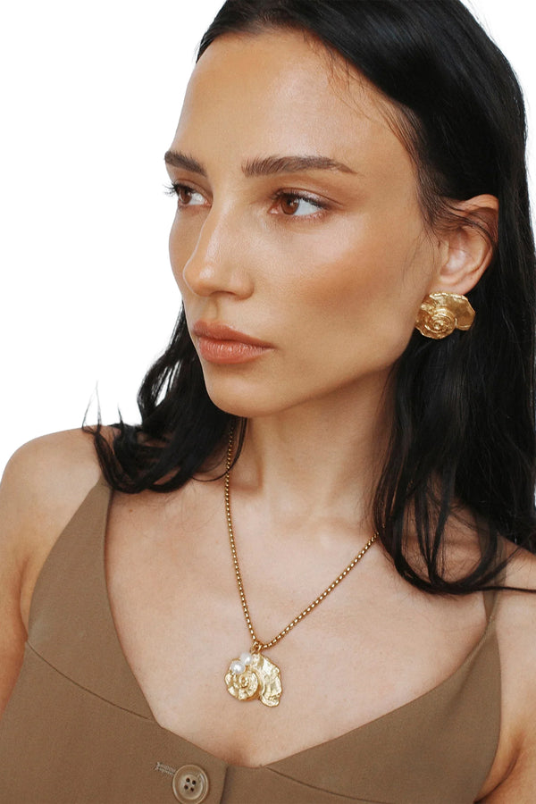 A close-up of a woman wearing Labelled Jewellery shell-shaped earrings in gold, paired with gold necklace and brown top, shown against a plaid white background.